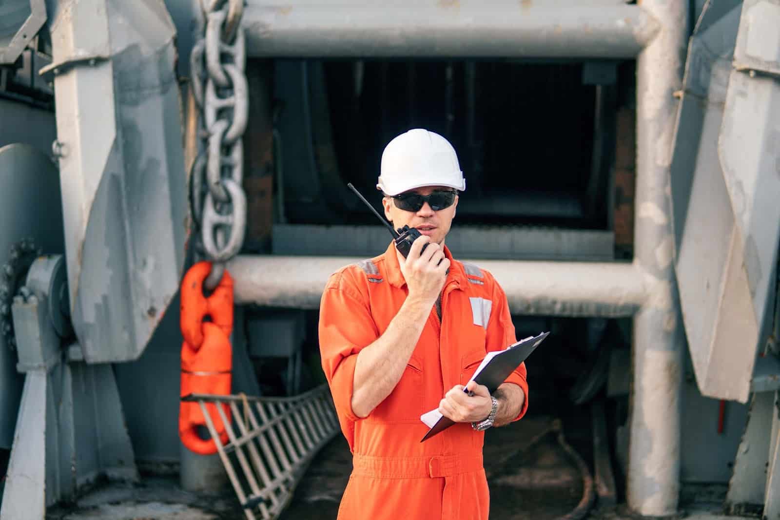 A worker in an orange coverall and white hard hat holds a clipboard and talks on a walkie-talkie, standing in front of industrial equipment—overseeing crew changes to assist crew managers with efficient operations.