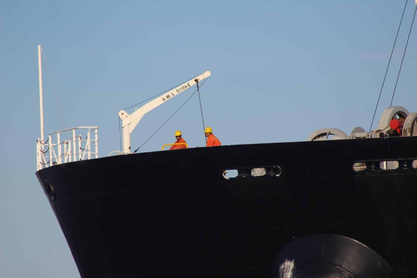 Two workers in orange uniforms and yellow helmets stand on the deck of a large black ship near a white crane, as crew managers oversee efficient crew changes against a clear blue sky.