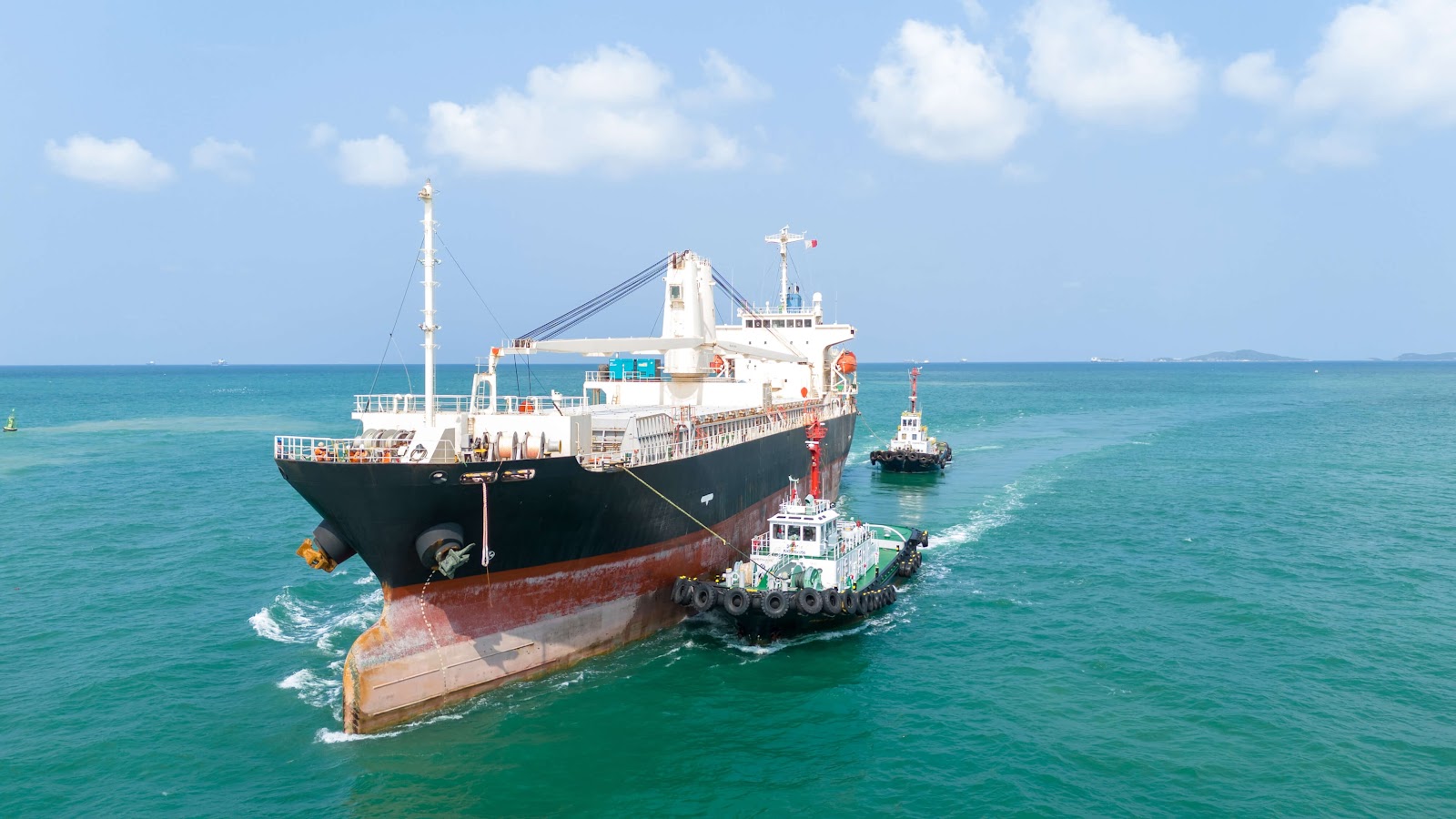 A large cargo ship, essential for smooth crew changes, is guided by two smaller tugboats on turquoise ocean water under a blue sky with scattered clouds.