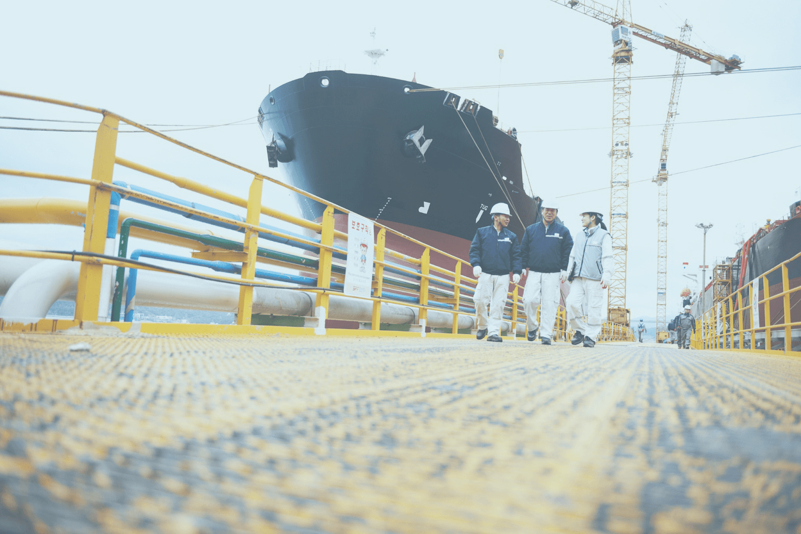 Four crew managers in safety helmets and jackets walk on a yellow metal platform near a large ship docked in a shipyard, with cranes and industrial piping, focusing on efficient crew changes in the busy port.