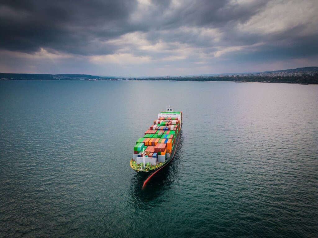 A large cargo ship loaded with colorful shipping containers sails across a calm body of water under a dramatic, cloudy sky, as port authorities monitor its voyage with distant land visible on the horizon.