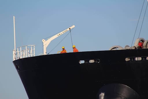 Two workers in orange uniforms and yellow helmets stand on the deck of a large black ship, near a white crane, preparing for ship to ship operation under a clear blue sky.