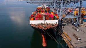 A large container ship docked at a port, stacked with colorful shipping containers, with cranes and vehicles for loading and unloading cargo visible on the right—a vital scene in the importer security filing deadline.