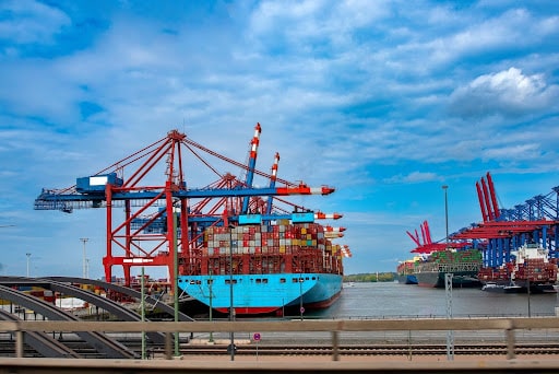 Large cargo ships loaded with colorful shipping containers are docked at a busy port, where importer security filing is essential. Red and blue cranes tower above as a road and rail tracks run in the foreground under a partly cloudy sky.