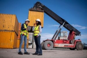 Two workers in safety vests and hard hats stand in front of stacked shipping containers, holding a clipboard and tablet with freight accounting info, with a large container handler vehicle behind them on a sunny day.