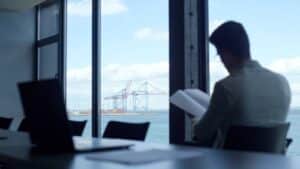 A person sits in an office by a window, reading documents related to maritime accounting. Outside, large shipping cranes and blue sky are visible across the water. A laptop sits on the table in the foreground.