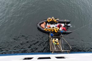 A group of people in yellow jackets sit in an inflatable boat next to a ship, as two crew members on a metal platform assist with a ship to ship personnel transfer over calm, dark water during sts operations.