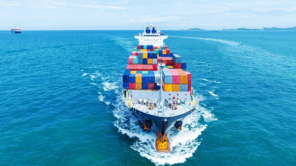 A cargo ship loaded with colorful shipping containers sails through bright blue ocean water under a partly cloudy sky—a vivid scene in the bustling maritime industry. Distant land and another ship are visible on the horizon.