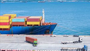 A large cargo ship loaded with colorful shipping containers is docked at the loading port during laycan. Trucks and cars are parked nearby on the concrete pier, and the blue sea is visible in the background.