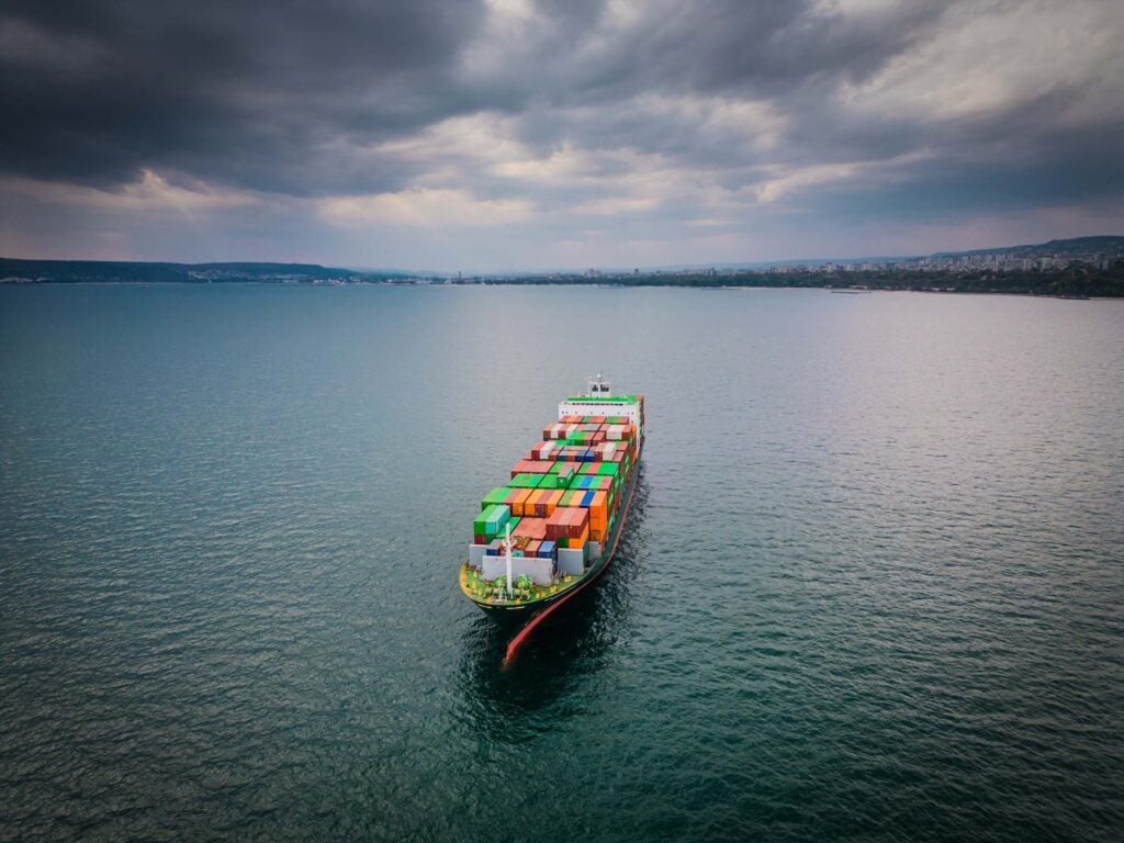 A large cargo ship carrying colorful shipping containers sails across a calm, open sea under a cloudy sky, illustrating the intricate shipping process as it heads toward a distant shoreline.