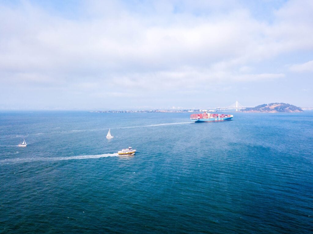 Aerial view of a large cargo ship highlighting the shipping process, alongside a ferry and two sailboats on blue water under a partly cloudy sky, with a distant coastline and bridge in the background.