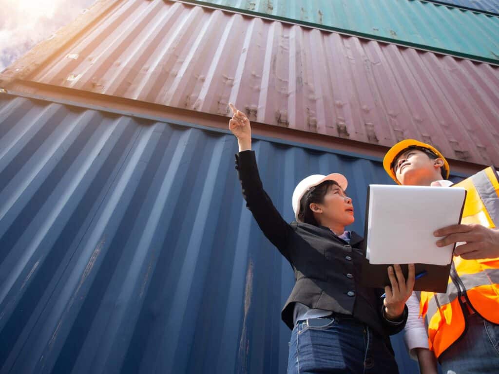 Two workers wearing safety helmets and vests stand before stacked shipping containers. One points upward while the other holds a clipboard, discussing the bill of lading vs packing list at a busy yard.