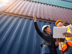 Two workers wearing safety helmets and vests stand before stacked shipping containers. One points upward while the other holds a clipboard, discussing the bill of lading vs packing list at a busy yard.
