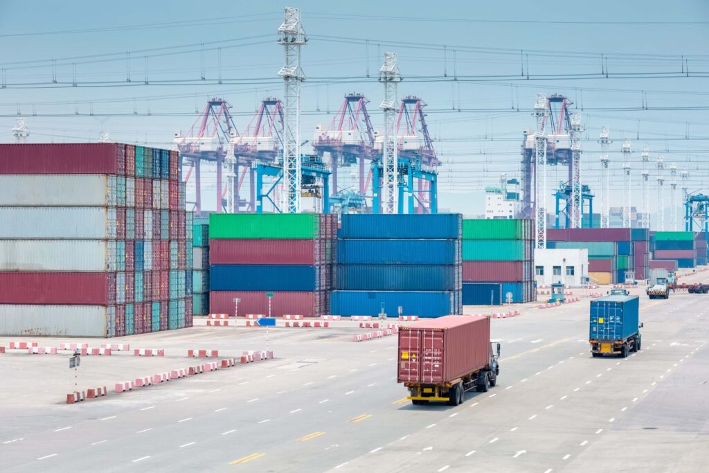 Shipping containers stacked in various colors at a busy port, with two trucks transporting containers on a wide road and large cranes visible in the background, as workers prepare for the next arrival notice.