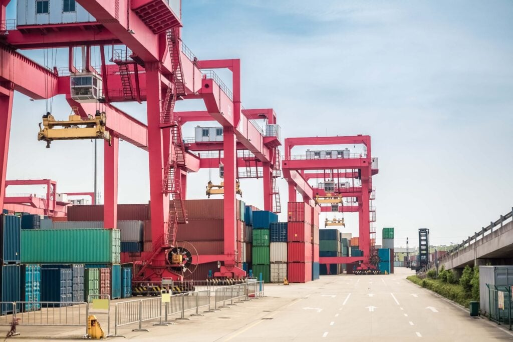 Rows of large red gantry cranes stand over stacks of colorful shipping containers at a shipping port, where an empty road runs alongside. The scene highlights efficient logistics processes like the automated manifest system (AMS filing).