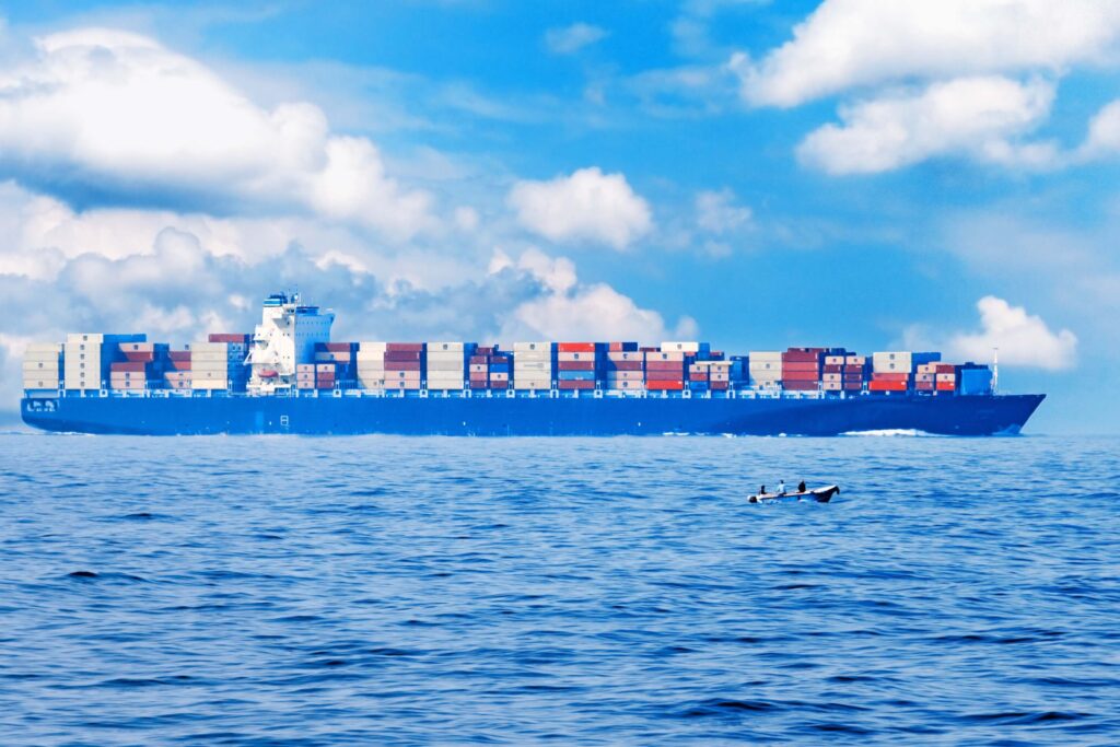 A large cargo ship loaded with colorful shipping containers, compliant with the automated manifest system, sails on a calm blue sea under a partly cloudy sky, with a small rowboat and two people in the foreground.