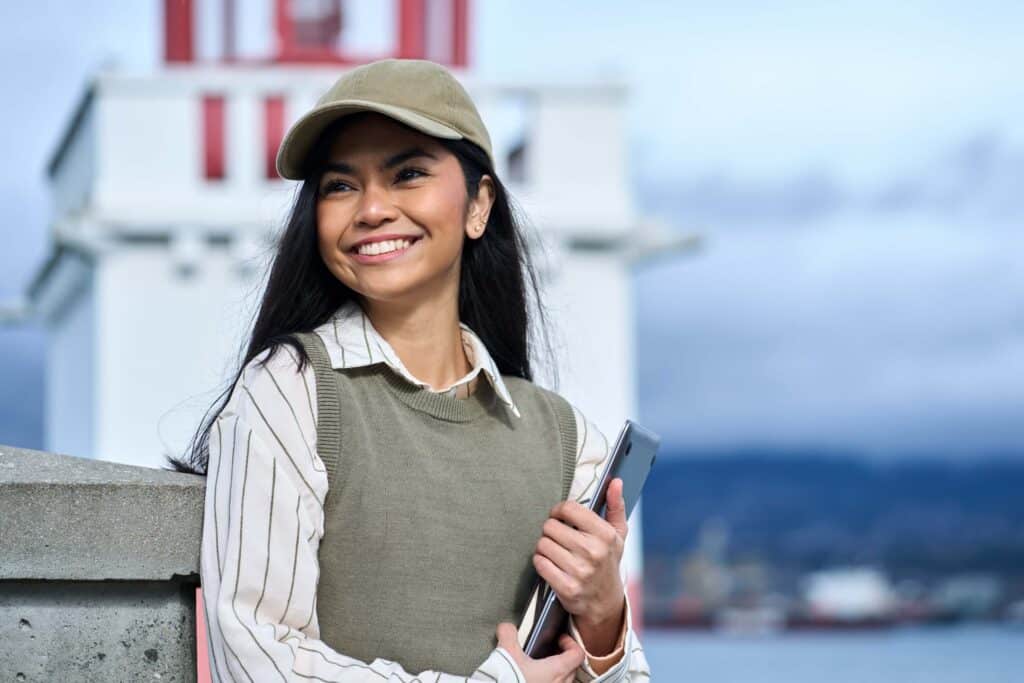 A young woman wearing a cap and sweater vest smiles while holding a tablet, standing outdoors in front of a white and red lighthouse, perhaps checking details related to the automated manifest system under a cloudy sky.