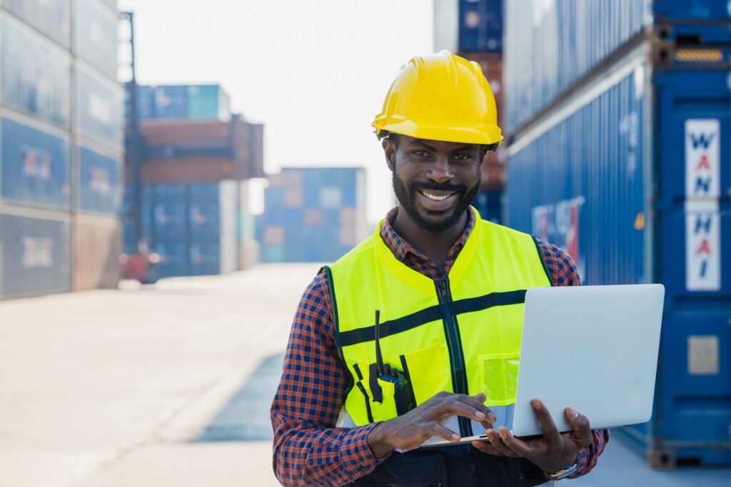 A smiling man, who is responsible for AMS filing, wears a yellow safety helmet and reflective vest as he stands outdoors at a shipping container yard, working on a laptop. Stacked shipping containers can be seen in the background.