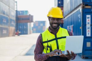 A smiling man, who is responsible for AMS filing, wears a yellow safety helmet and reflective vest as he stands outdoors at a shipping container yard, working on a laptop. Stacked shipping containers can be seen in the background.