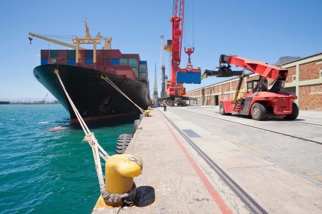 A large cargo ship docked at a port with shipping containers being loaded by a red crane and a reach stacker, as workers verify the arrival date under a clear blue sky.