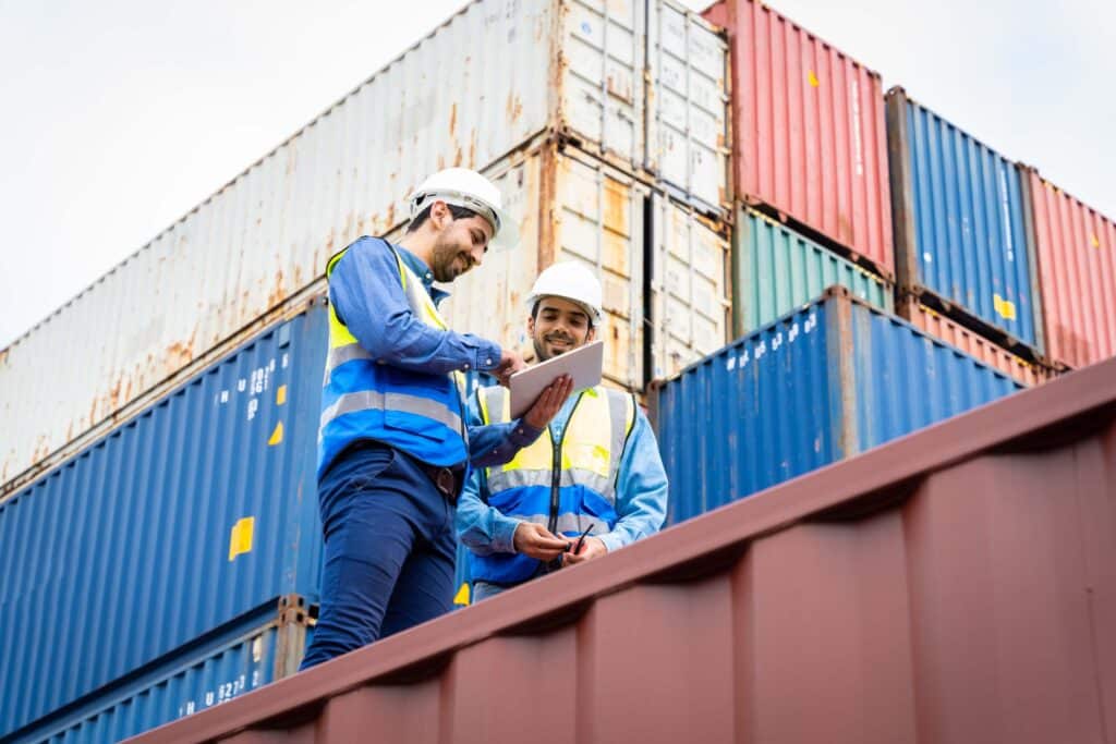 Two workers wearing safety helmets and reflective vests check a clipboard—likely reviewing a packing slip—while standing by colorful shipping containers in a freight yard.