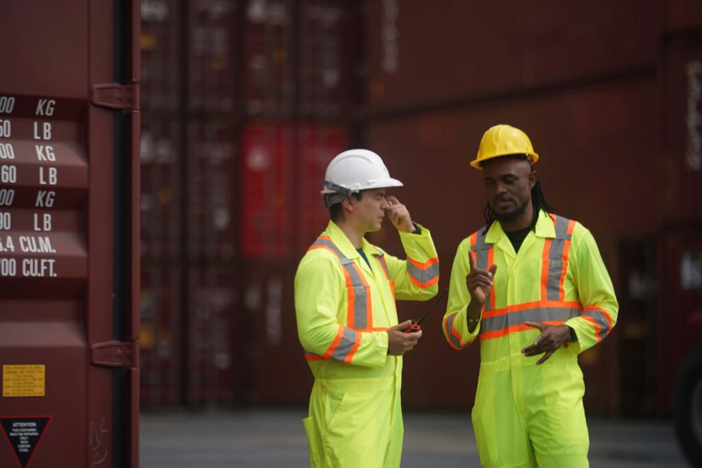 Two workers in yellow high-visibility safety suits and hard hats stand by large shipping containers, discussing maritime automated invoice processing as one gestures and the other adjusts their helmet.