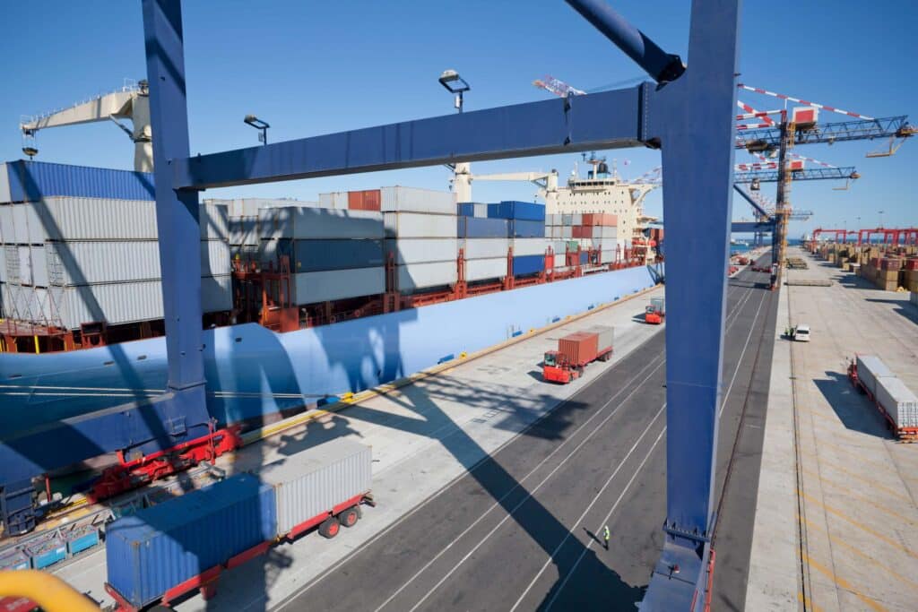 A large cargo ship docked at a busy port is surrounded by cranes moving containers, while trucks wait along the pier under a clear blue sky—an efficient scene of port call management in action.