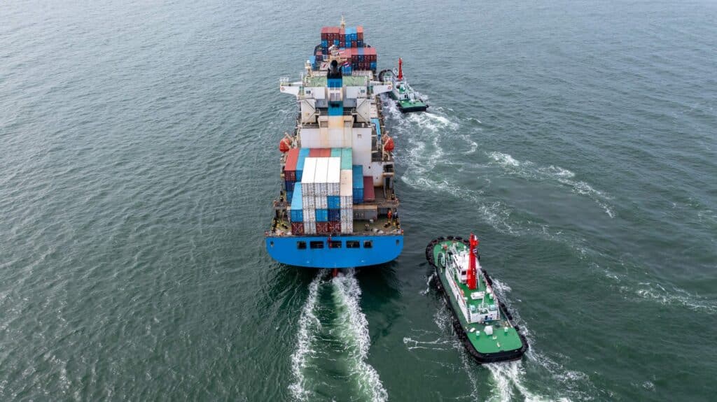 Aerial view of a large cargo ship carrying colorful containers at sea, escorted by two smaller tugboats during cargo operations, creating white wakes in the water.