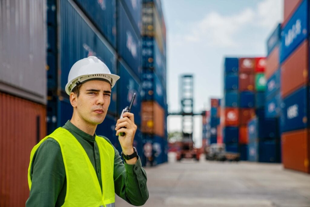 A man wearing a white hard hat and yellow safety vest holds a walkie-talkie, standing in a shipping container yard with stacked containers and industrial equipment, overseeing port call documentation.