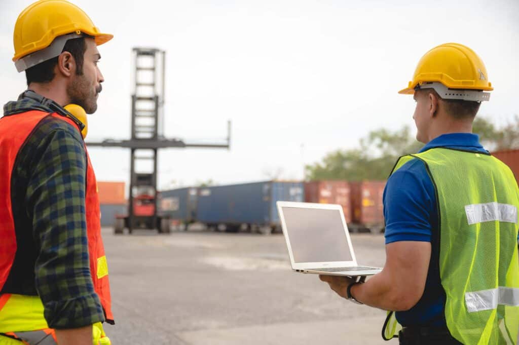 Two workers in safety vests and yellow helmets stand outside near shipping containers; one holds a laptop as they observe a container crane, likely discussing port call management and overseeing efficient port calls.