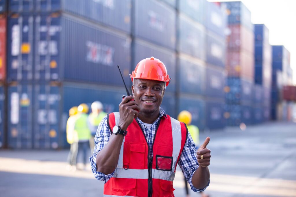A smiling worker in a red safety vest and hard hat holds a walkie-talkie, giving a thumbs up at a shipping container yard, ready to issue a notice of readiness as other workers and stacked containers fill the background.