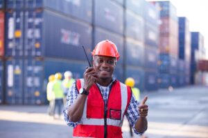 A smiling worker in a red safety vest and hard hat holds a walkie-talkie, giving a thumbs up at a shipping container yard, ready to issue a notice of readiness as other workers and stacked containers fill the background.