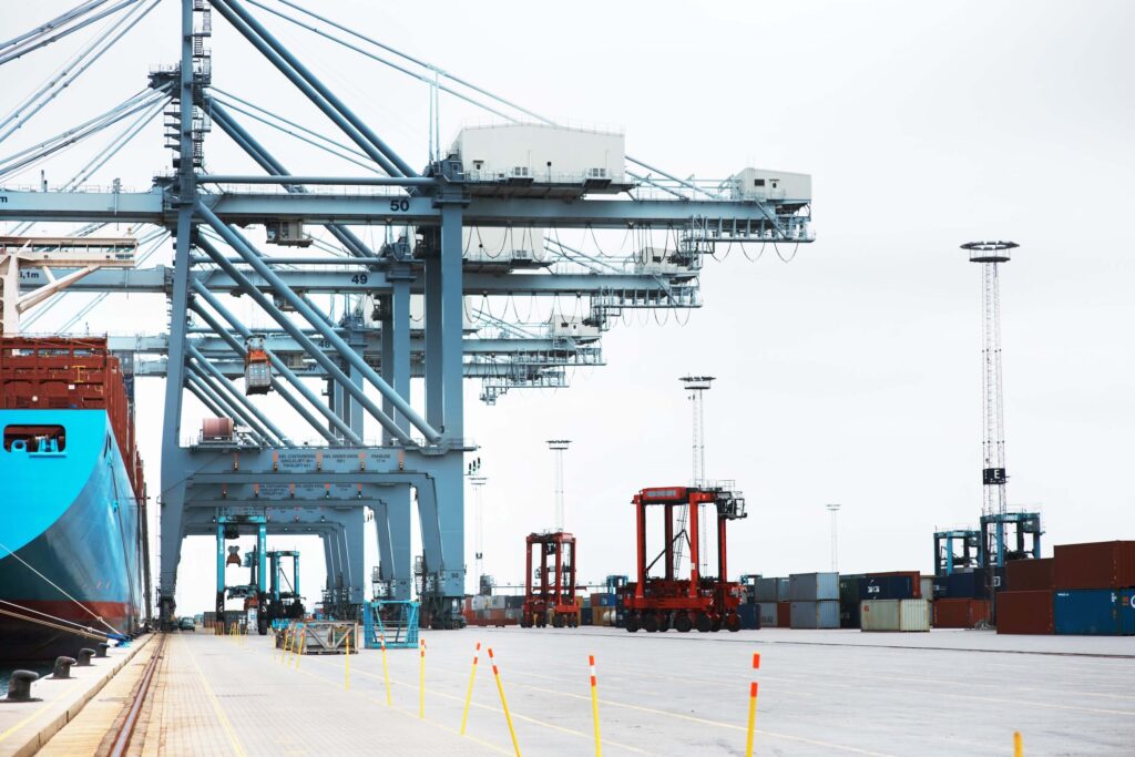 Large shipping cranes and red cargo handling vehicles operate beside stacked shipping containers at a busy port, with a docked container ship partially visible on the left. The scene is industrial and expansive.