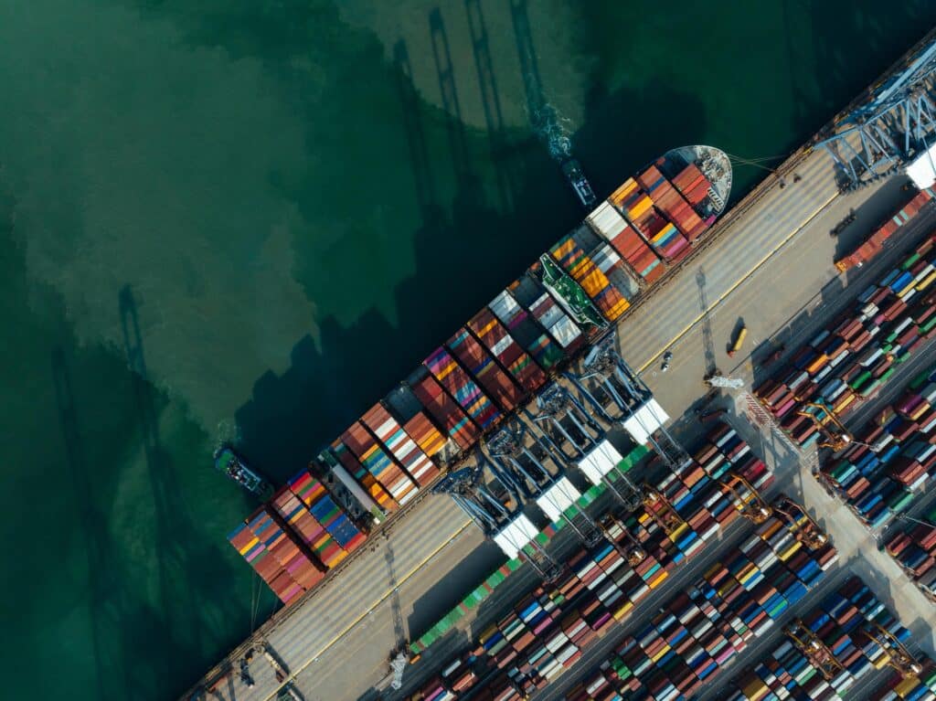 Aerial view of a busy shipping port with a large cargo ship docked, loaded with colorful containers. Cranes are visible unloading the containers, and the water appears deep green beside the dock.