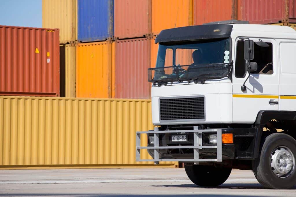 A white truck is parked in front of a stack of colorful shipping containers, highlighting the busy atmosphere typical of a freight RFQ or procurement process at a shipping yard or industrial area.