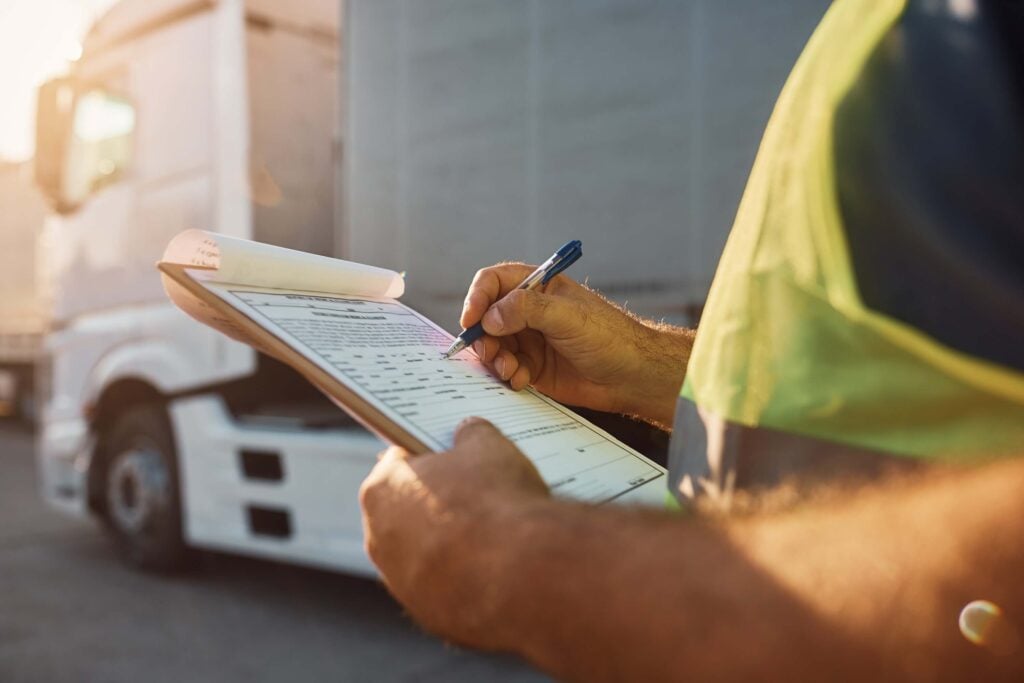 A person in a high-visibility vest writes on a clipboard, reviewing freight RFQ documents next to a white truck in an outdoor industrial setting.