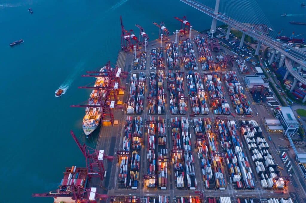 Aerial view of a busy shipping port with rows of colorful cargo containers, cranes loading and unloading ships, and a boat moving through blue water near a large bridge.