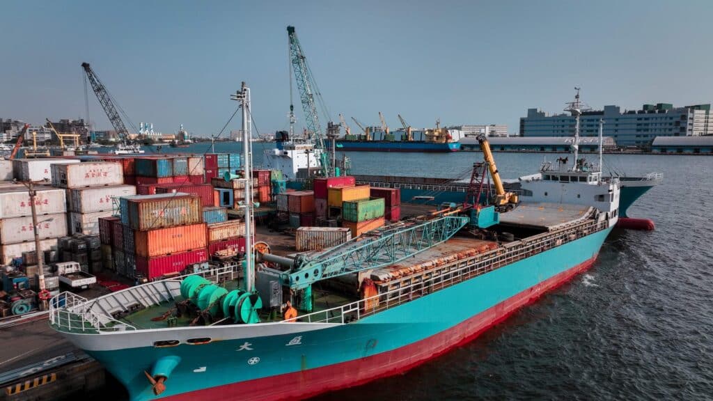 A large cargo ship docked at a busy port during a port call handover, surrounded by colorful stacked shipping containers and cranes, with buildings and more ships visible in the background under a clear sky.