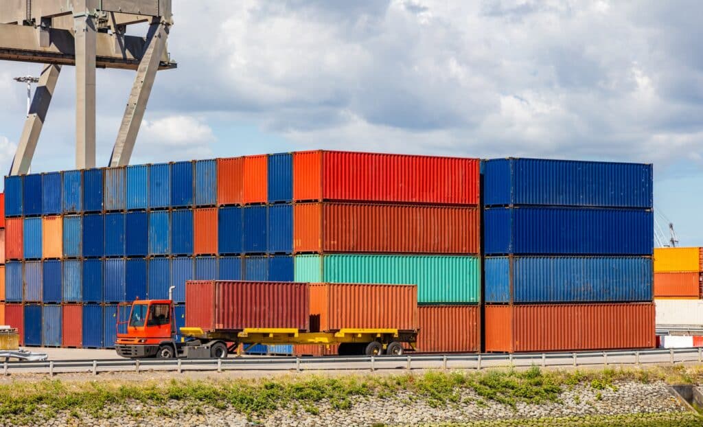 A truck transports a shipping container in front of large stacks of colorful cargo containers at a shipping yard, showcasing the scale managed by logistics service providers, with a partly cloudy sky in the background.
