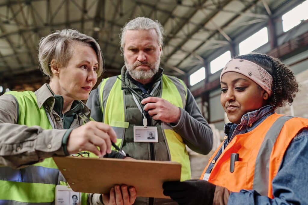 Three workers wearing safety vests and ID badges stand together in a warehouse. One woman is holding a clipboard and pointing, while the other two listen attentively. The setting appears industrial with a high ceiling.