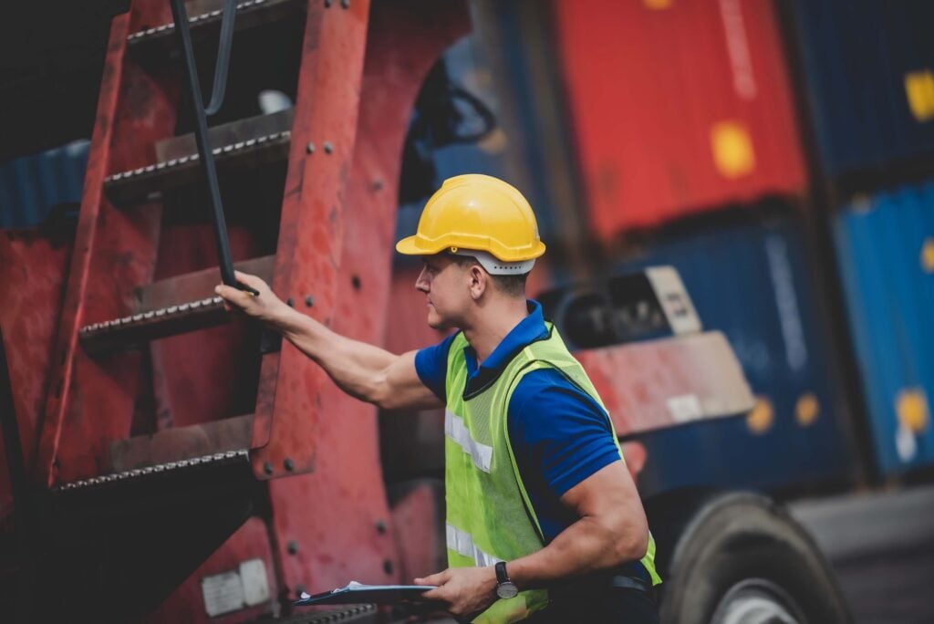 A worker wearing a yellow hard hat, green safety vest, and blue shirt holds onto a red industrial ladder at a shipping yard, where stacked cargo containers highlight the busy freight RFQ and procurement process.