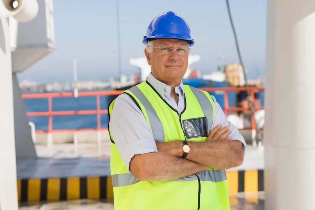 An older man wearing a blue hard hat, glasses, and a reflective safety vest stands with arms crossed on an industrial site near water, possibly overseeing a draft survey, with industrial equipment and fencing in the background.