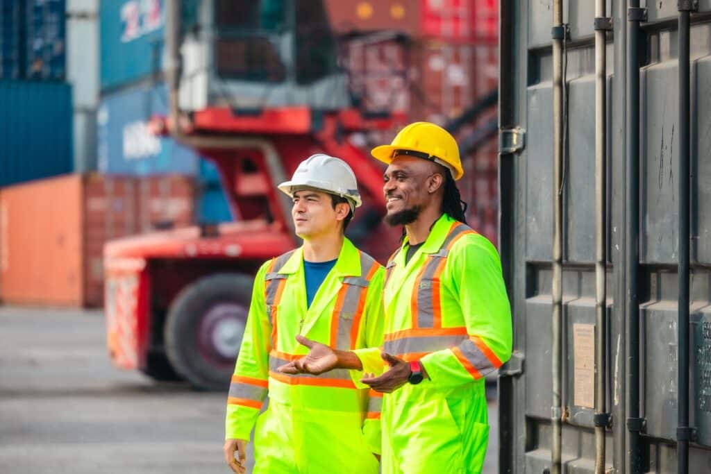 Two workers wearing neon yellow safety jackets and hard hats stand outside near shipping containers and heavy machinery, smiling as they oversee the port call process.