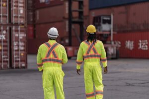 Two workers in yellow high-visibility suits and hard hats walk side by side in a shipping yard, one holding the mate's receipt, with stacked cargo containers in the background.