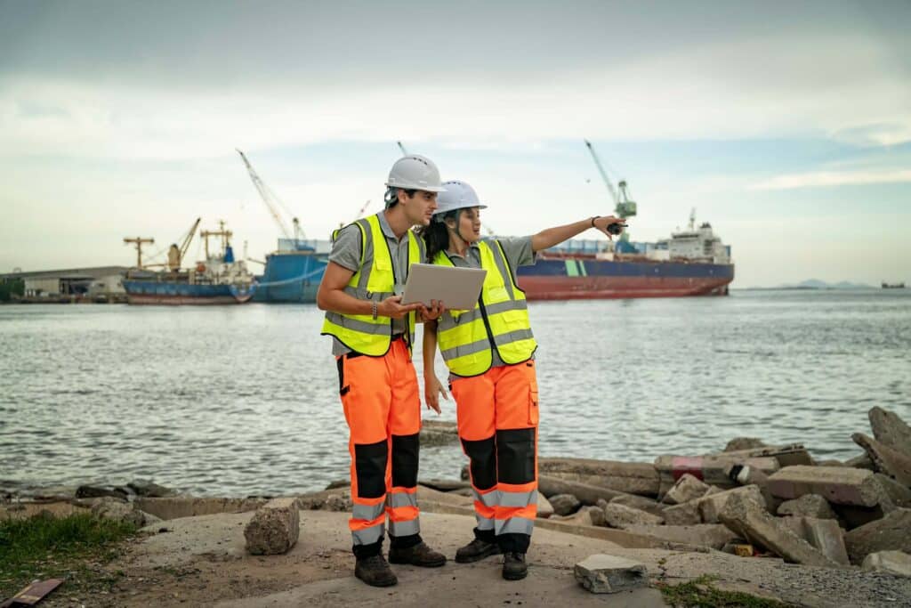 Two construction workers in safety gear and helmets stand by the water with ships and cranes in the background; one holds a laptop displaying port data while the other points toward the harbor.
