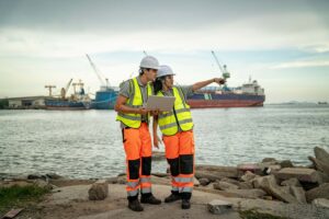 Two construction workers in safety gear and helmets stand by the water with ships and cranes in the background; one holds a laptop displaying port data while the other points toward the harbor.