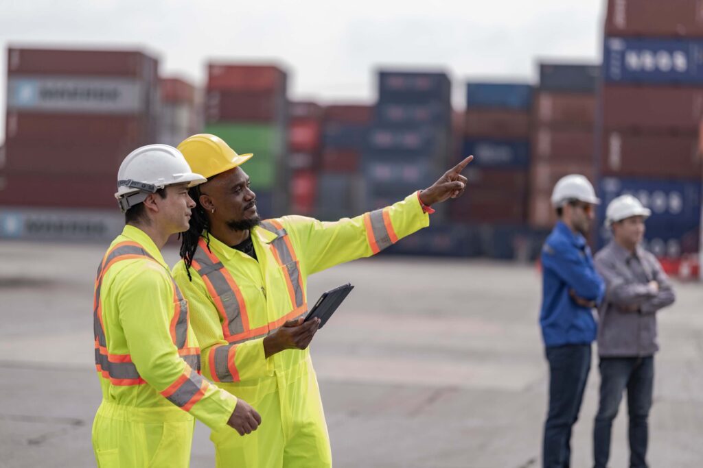 Two workers in yellow safety uniforms and helmets stand at a shipping yard; one points ahead while holding a tablet. Stacks of shipping containers and two other workers are visible in the background.