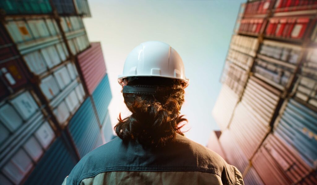A person wearing a white hard hat stands between tall stacks of shipping containers, looking up at them under a bright sky, perhaps assessing cargowise pricing for logistics operations.