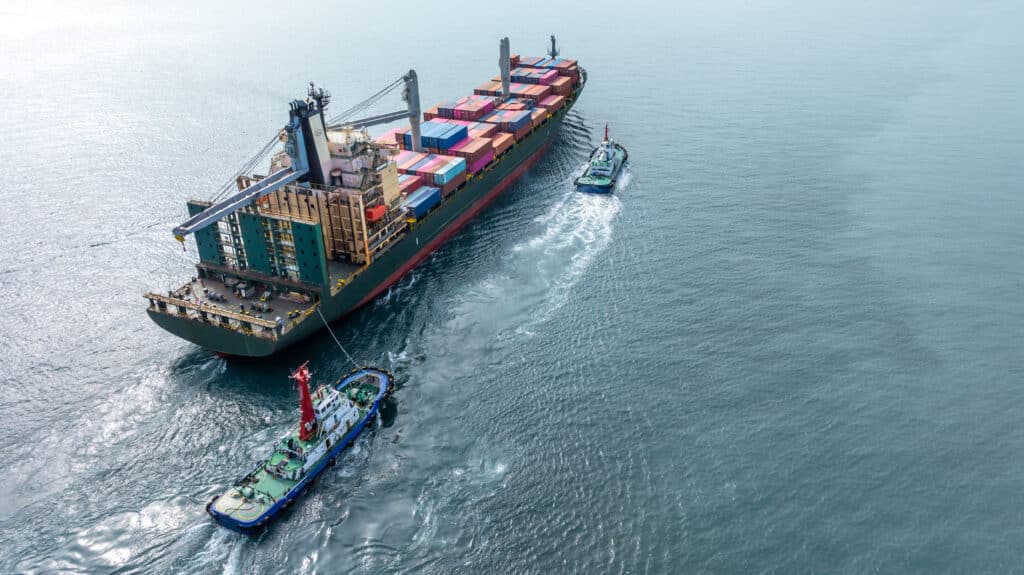 Aerial view of a large cargo ship carrying colorful shipping containers being guided by two tugboats across calm, open water, incurring a total pilotage fee as part of its safe passage.