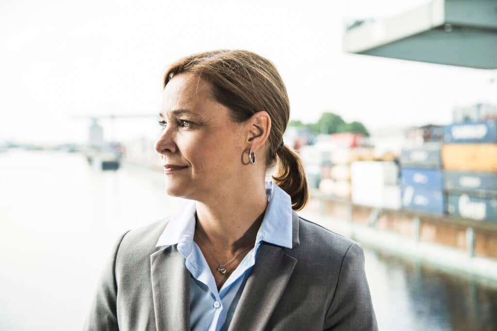 A woman in a gray blazer and blue collared shirt stands by a waterfront, looking to the side. Shipping containers and industrial structures—common sights near pilotage charge zones—are visible in the blurred background.